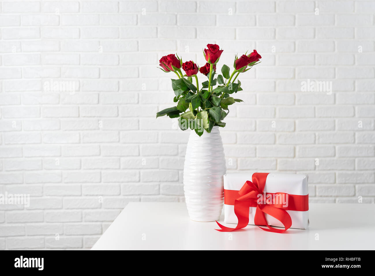 Red roses in white vase next to a gift box with red ribbon against white brocks wall Stock Photo