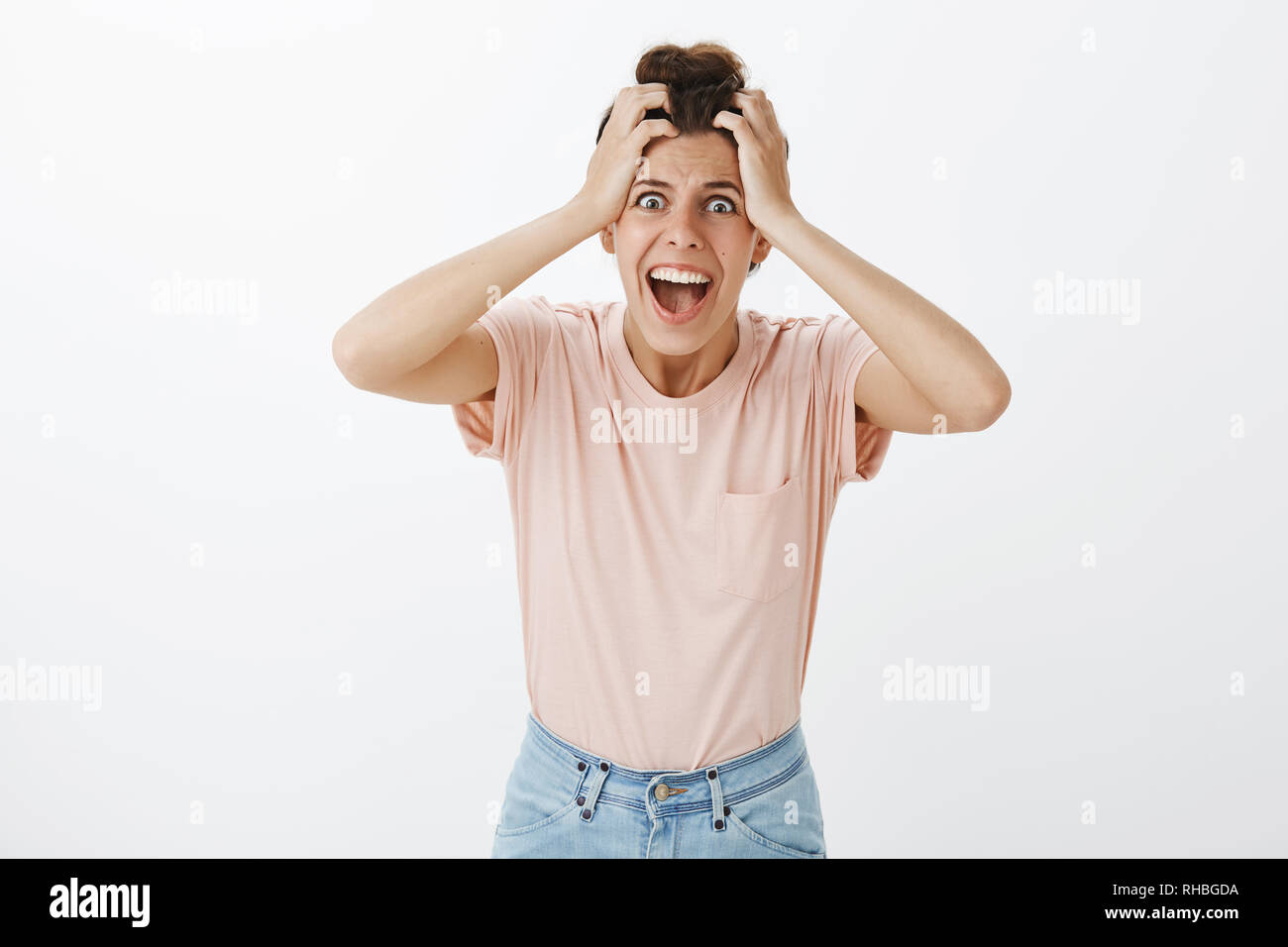 Freaked out concerned and shouting young insane girlfriend with messy bun holding hand on head yelling at camera with popped eyes losing temper as Stock Photo
