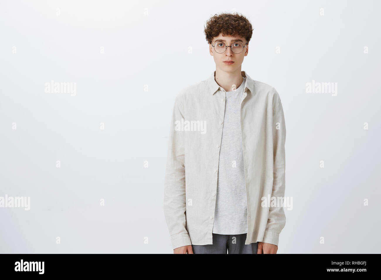 Studio shot of indifferent unemotional handsome and stylish european hipster boy in glasses with curly haircut standing upright looking at camera with Stock Photo