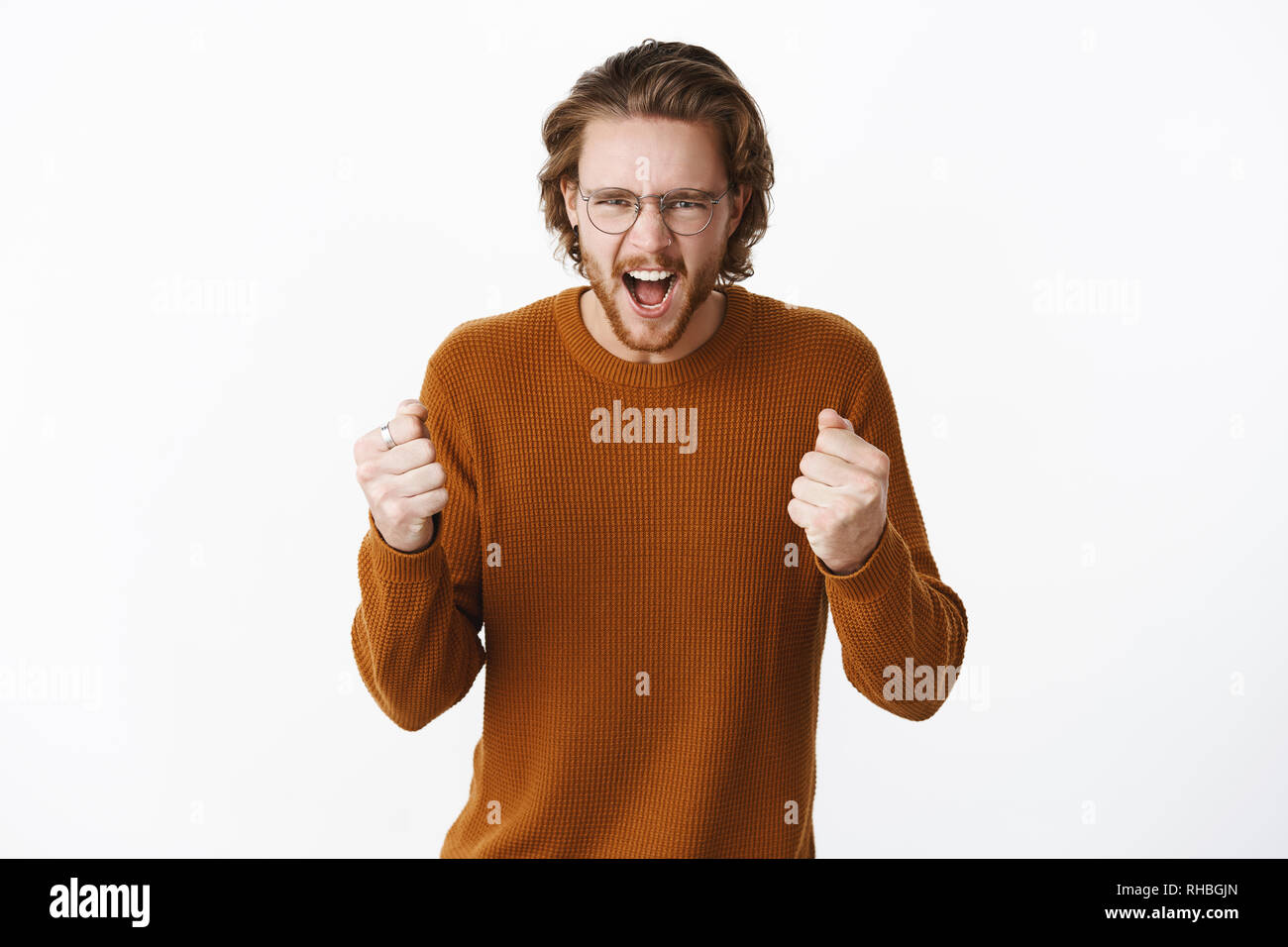 Studio shot of expressive good-looking european bearded guy in sweater and glasses yelling with fure and rage clenching raised fists as cheering and Stock Photo