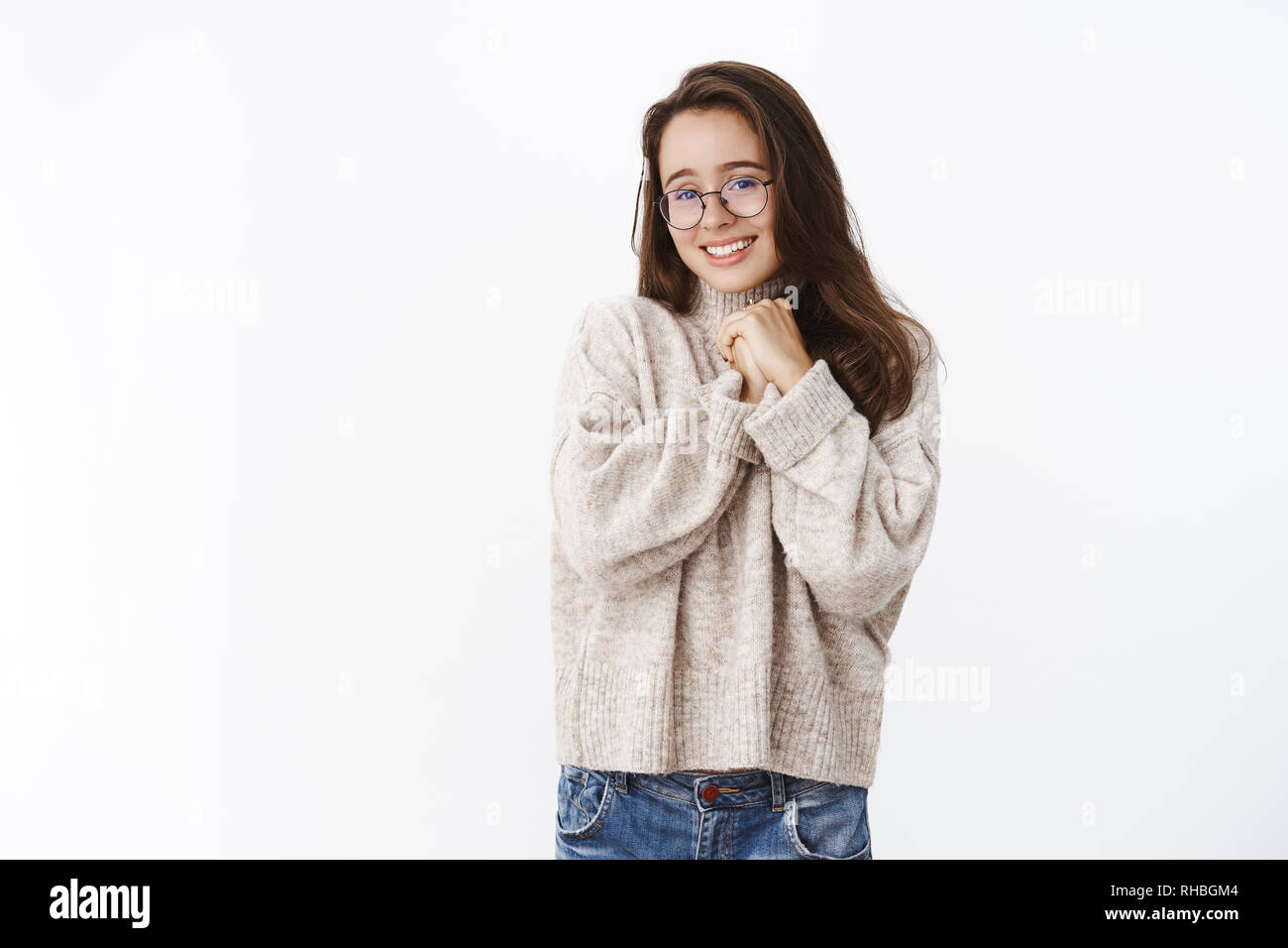 Silly charming girlfriend in glasses and sweater holding hands together pressed to chest and smiling with love, tender feelings, being in love Stock Photo