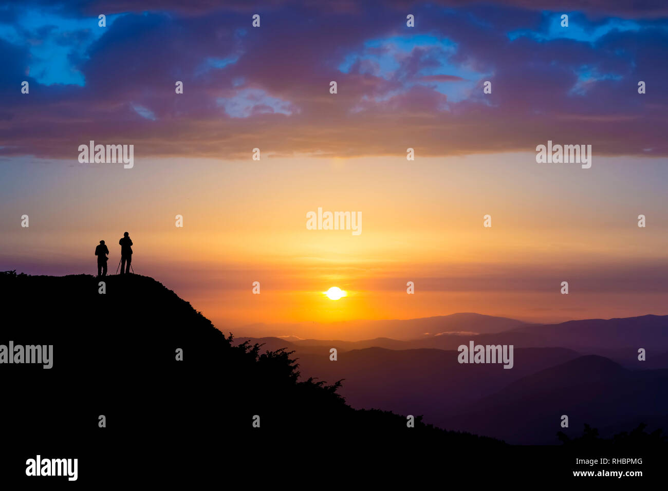 Silhouettes of two people standing on a rock and looking toward the sun. Dramatic clouds over them. Sunset in the mountains. Purple light. Summer in t Stock Photo