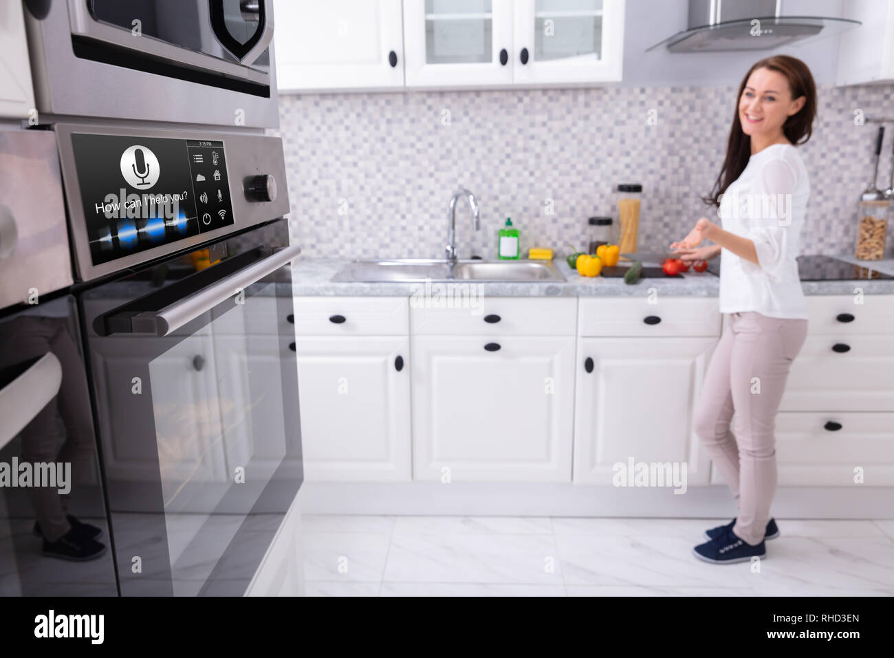 Happy Young Woman Looking At Oven With Voice Recognition Function In Kitchen Stock Photo