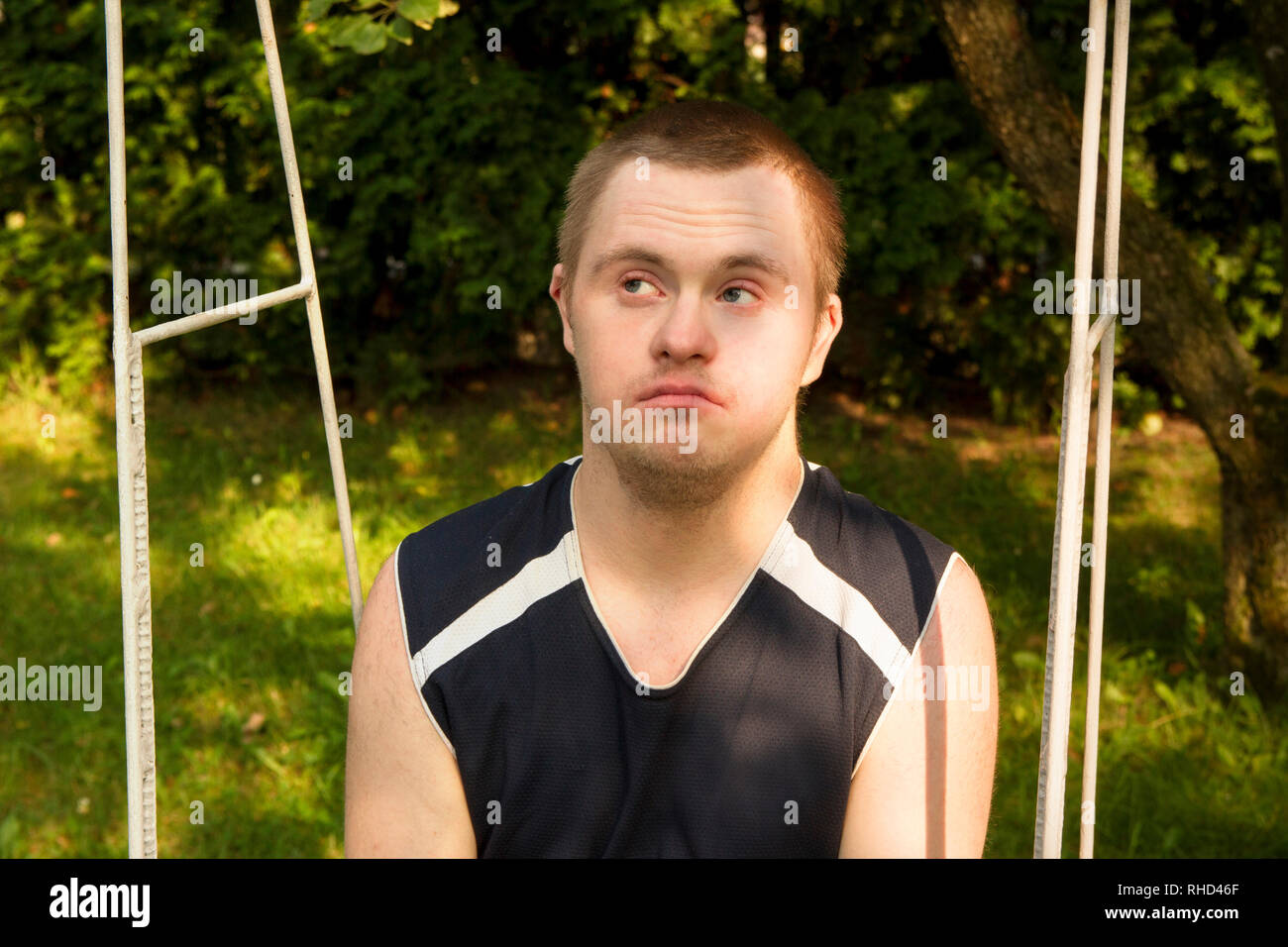 Portrait of young person with Down Syndrome sitting on a swing in the garden, backyard during summer day. Young handsome looks up looks up. Face close Stock Photo