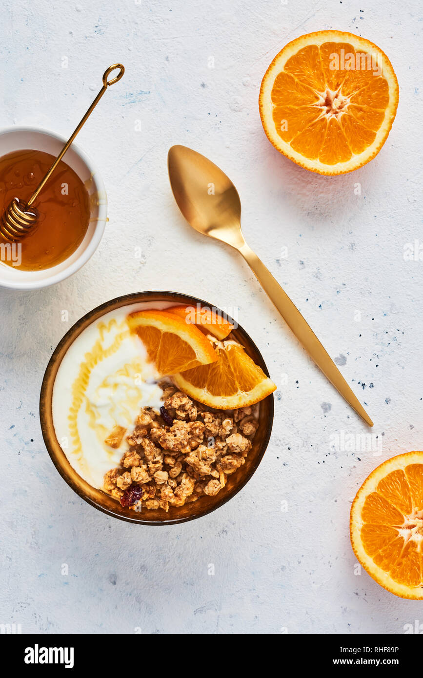 Bowl of homemade granola with yogurt, honey and fresh orange on white abstract background. Healthy breakfast. Golden utensils. Top view. Copy space fo Stock Photo