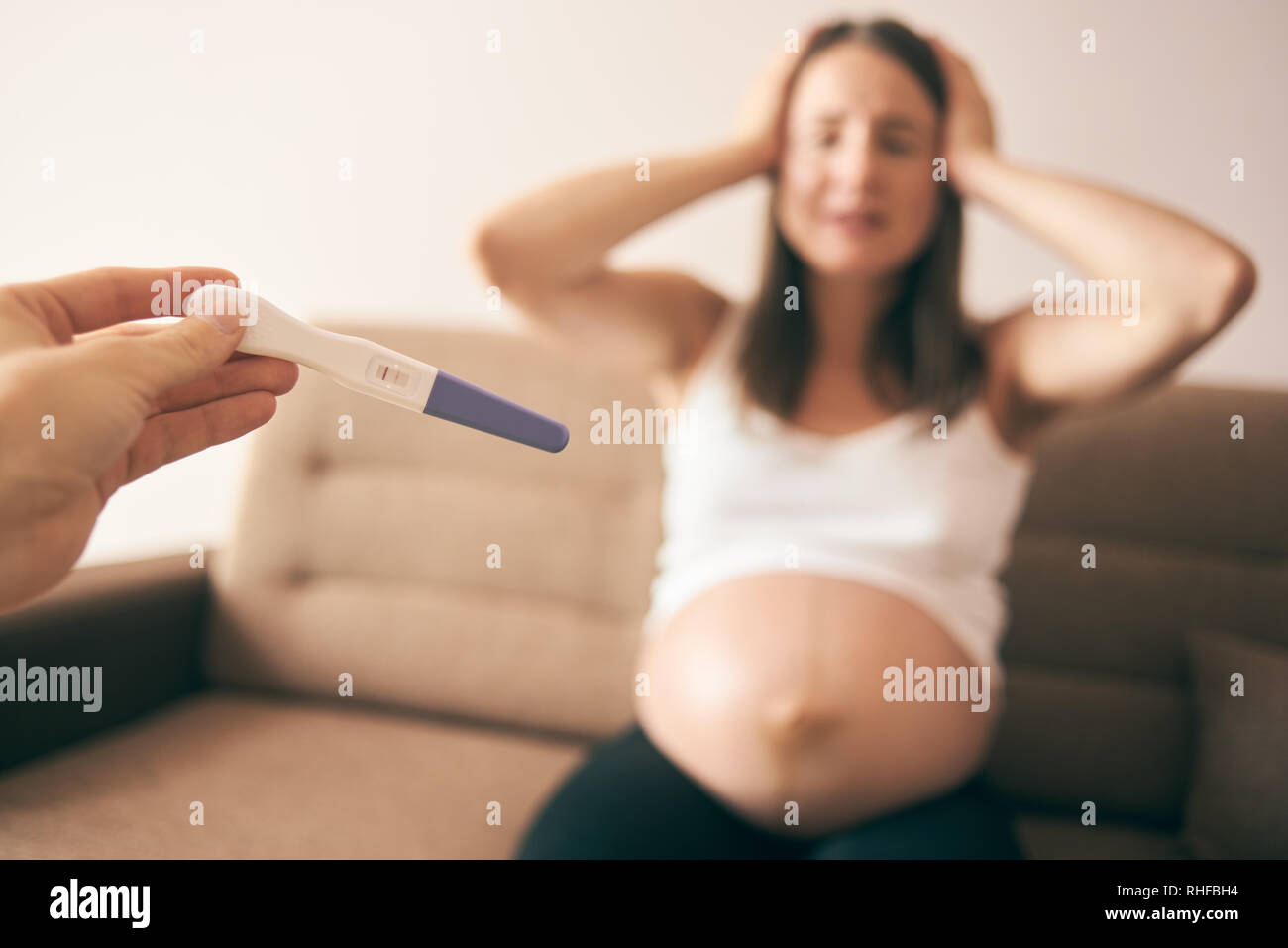 Selective focus of pregnancy test on male hand. Sad pregnant woman in white shirt and in despair and crying sitting on sofa at background. Concept of disappointment and unwanted child. Stock Photo