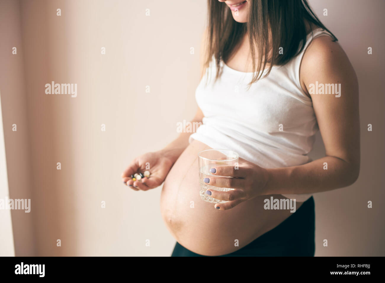 Side view of smiling pregnant woman with big belly standing at home and taking vitamins with water. Young future mother caring about health. Concept of expectation and parental love. Stock Photo