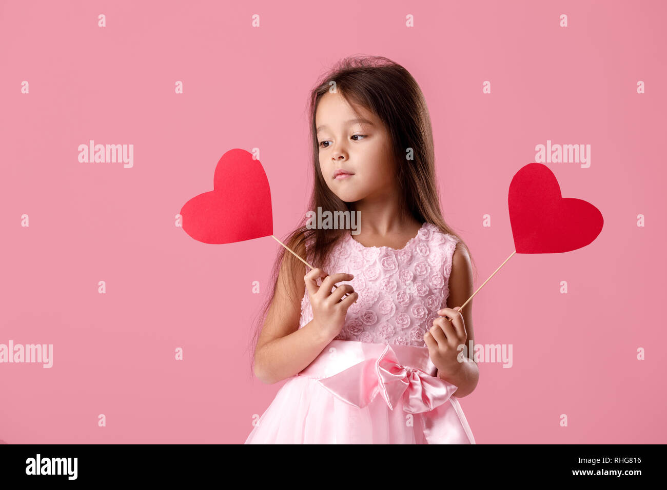 portrait of a cute little girl in a pink dress holding two paper heart ...