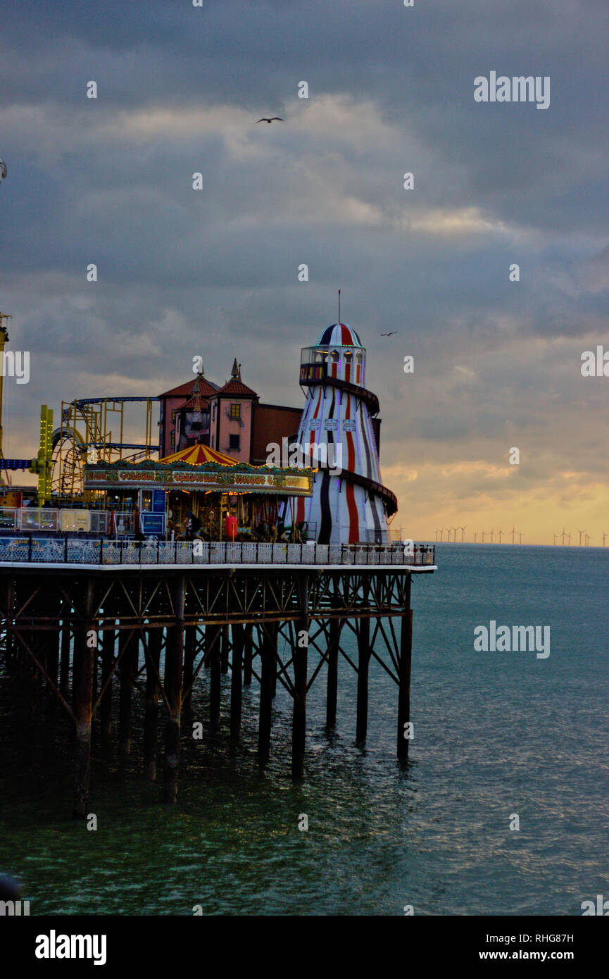 Slide, carousel and other fair attractions on the platform of Brighton Pier. Cloudy sky with warm sunlight in the background. Stock Photo