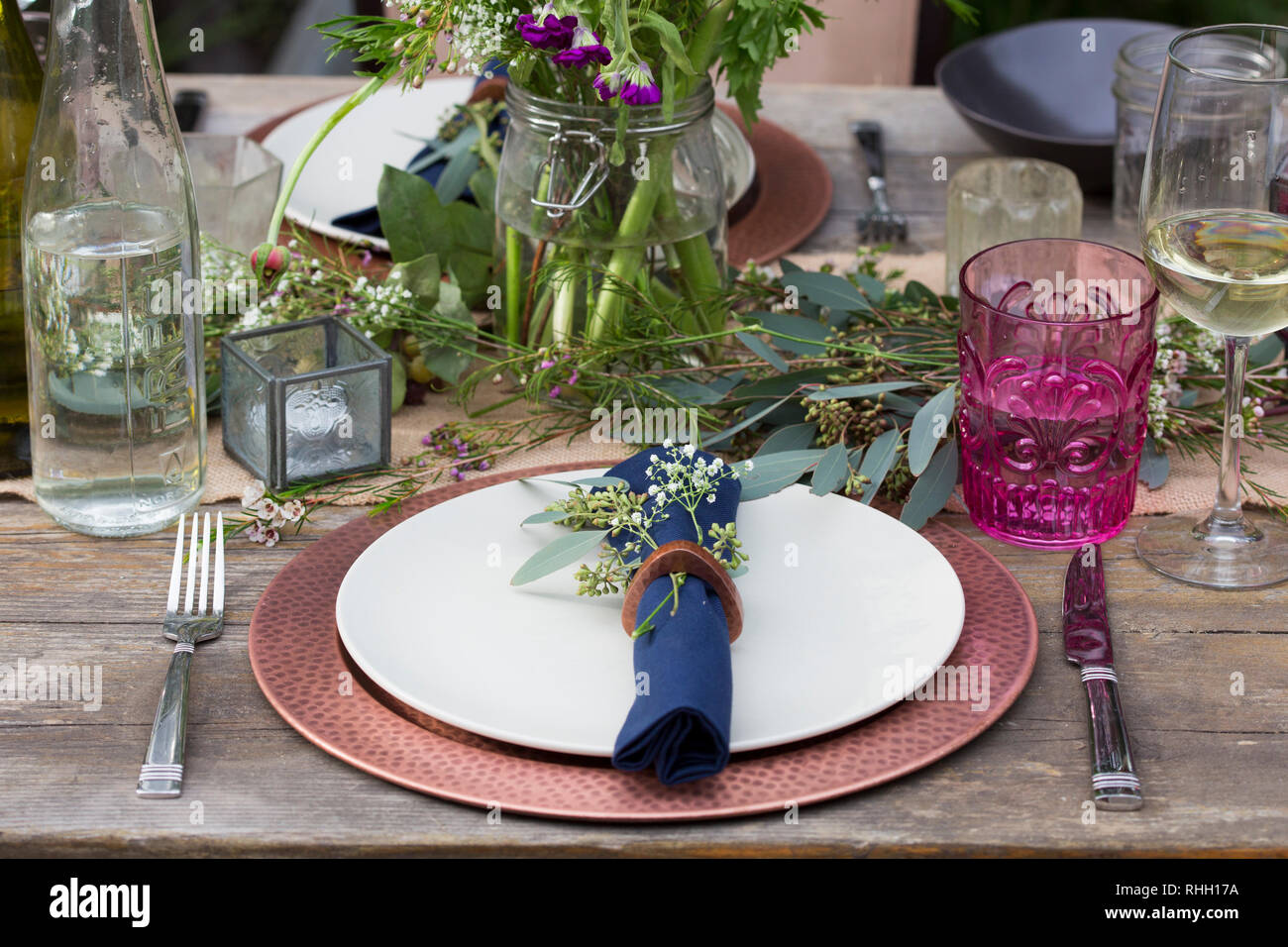 Blue napkin in ring on white plate with copper charger, place setting on wooden outdoor dinner party dining table with flowers, glass, and candle. Stock Photo