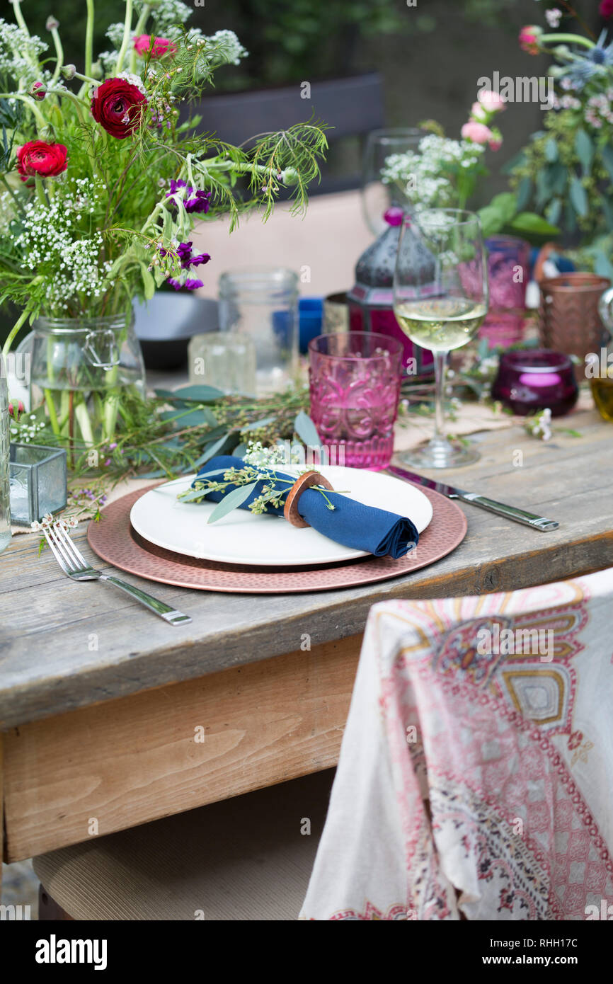 Blue napkin in ring on white plate with copper charger, place setting on wooden outdoor dinner party dining table with flowers, glass, and candle. Stock Photo
