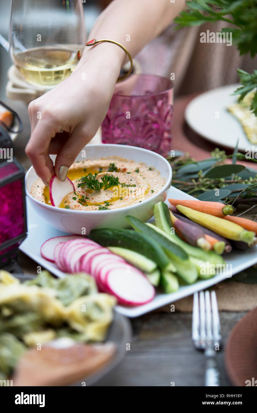 Female hands dipping radish in bowl of hummus at outdoor dinner party. Stock Photo