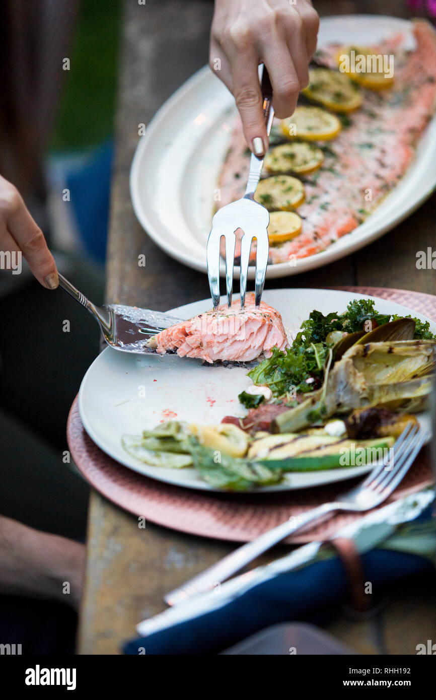 Hands serving salmon with lemon with silver fork from platter to plate with vegetables. Stock Photo
