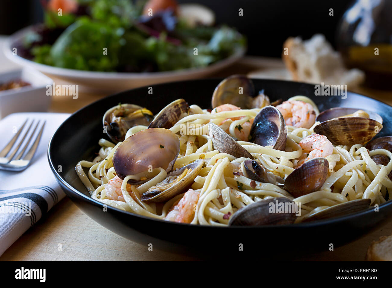 Bowl of spaghetti pasta with seafood- clams and shrimp- on wooden table with fork, napkin, and side salad. Stock Photo