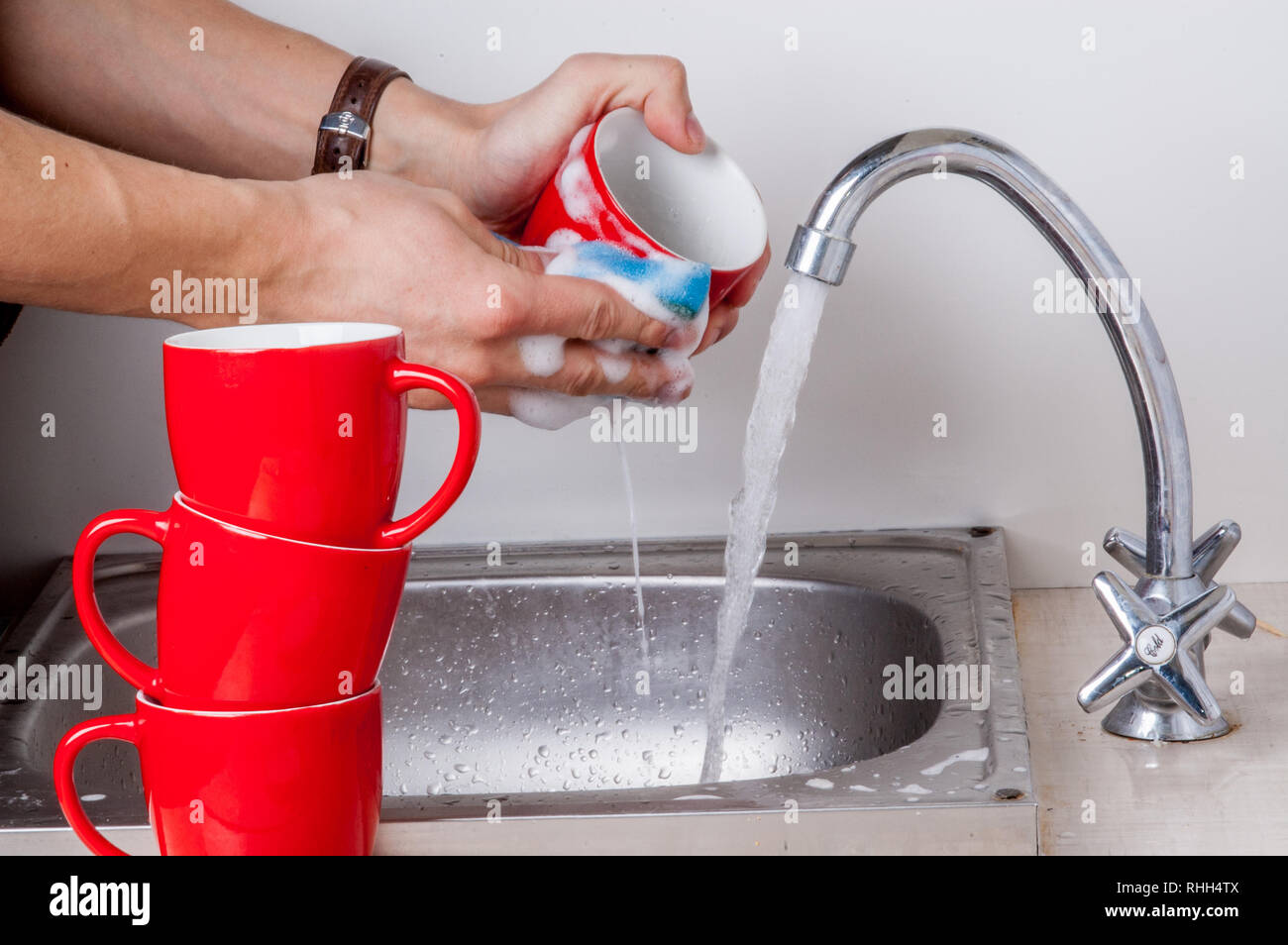 Male hands are washing red cups with sponge under running water in the kitchen sink Stock Photo