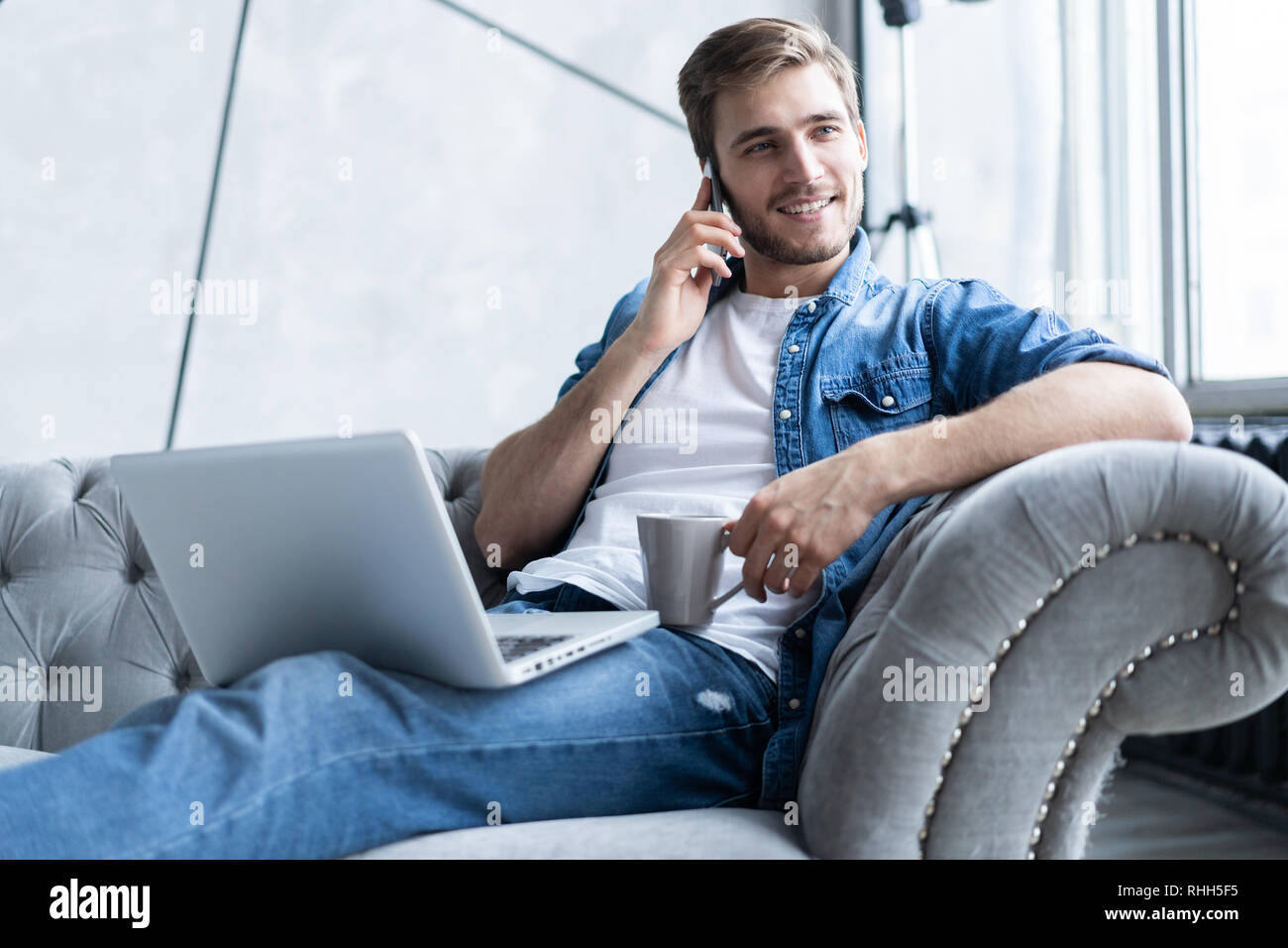 Portrait of handsome young man making a call and using his laptop while sitting on sofa at home Stock Photo