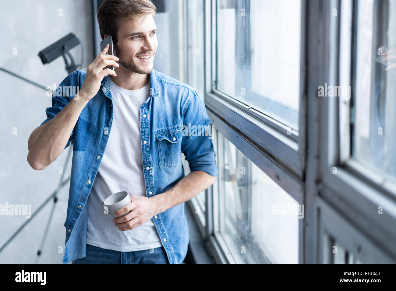 Smiling young man talking on mobile phone, looking at the window at home. Stock Photo