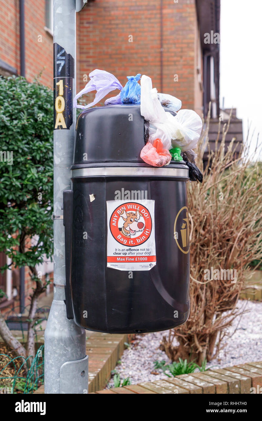 Overflowing black plastic waste bin attached to a metal post with dog waste information sticker Stock Photo