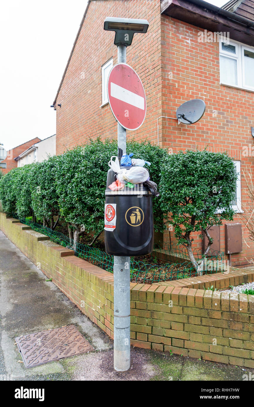 Overflowing black plastic waste bin attached to a metal post below a no entry road sign Stock Photo