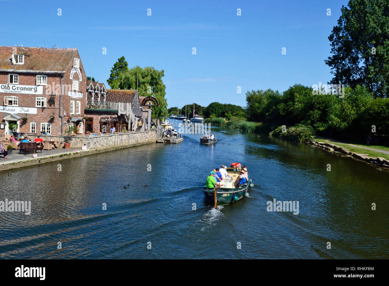 People on a small motor boat travelling along The River Frome. Buildings at The Quay, Wareham in Dorset UK, include The Old Granary, cafe and bar. Stock Photo