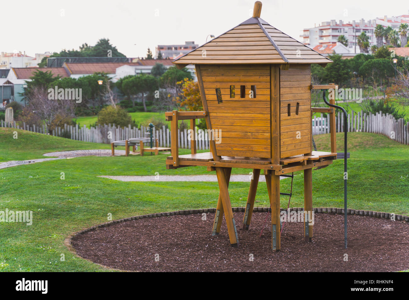 Yellow brown wooden tree house, kids playground equipment at park with green grass, bushes, trees, and a wooden fence, Oeiras Portugal Stock Photo