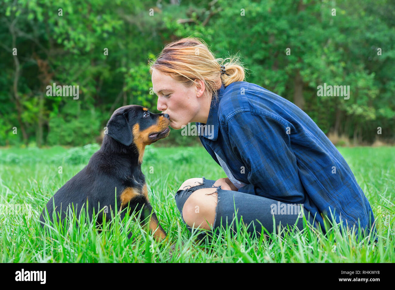 Young caucasian female dog owner  kisses rottweiler puppy on head in natural meadow Stock Photo