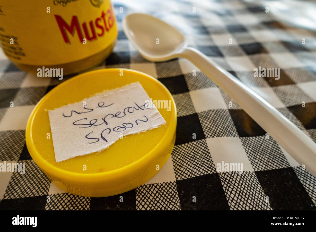 Closeup of Colmans English Mustard jar with lid off and sticker saying use seperate spoon. plastic spoon on a black and white plastic gingham table cl Stock Photo