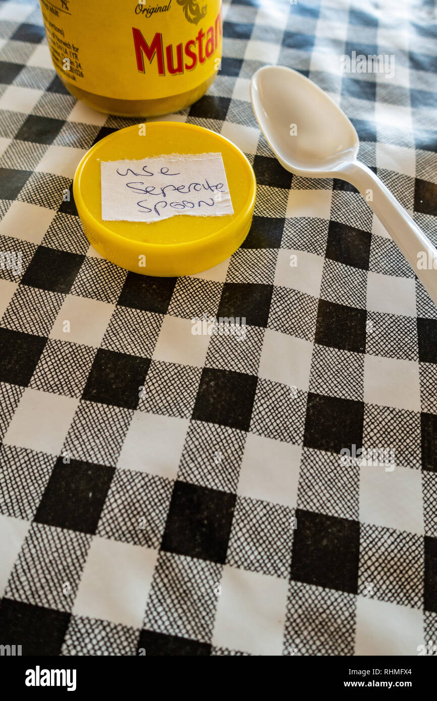 Closeup of Colmans English Mustard jar with lid off and sticker saying use seperate spoon. plastic spoon on a black and white plastic gingham table cl Stock Photo