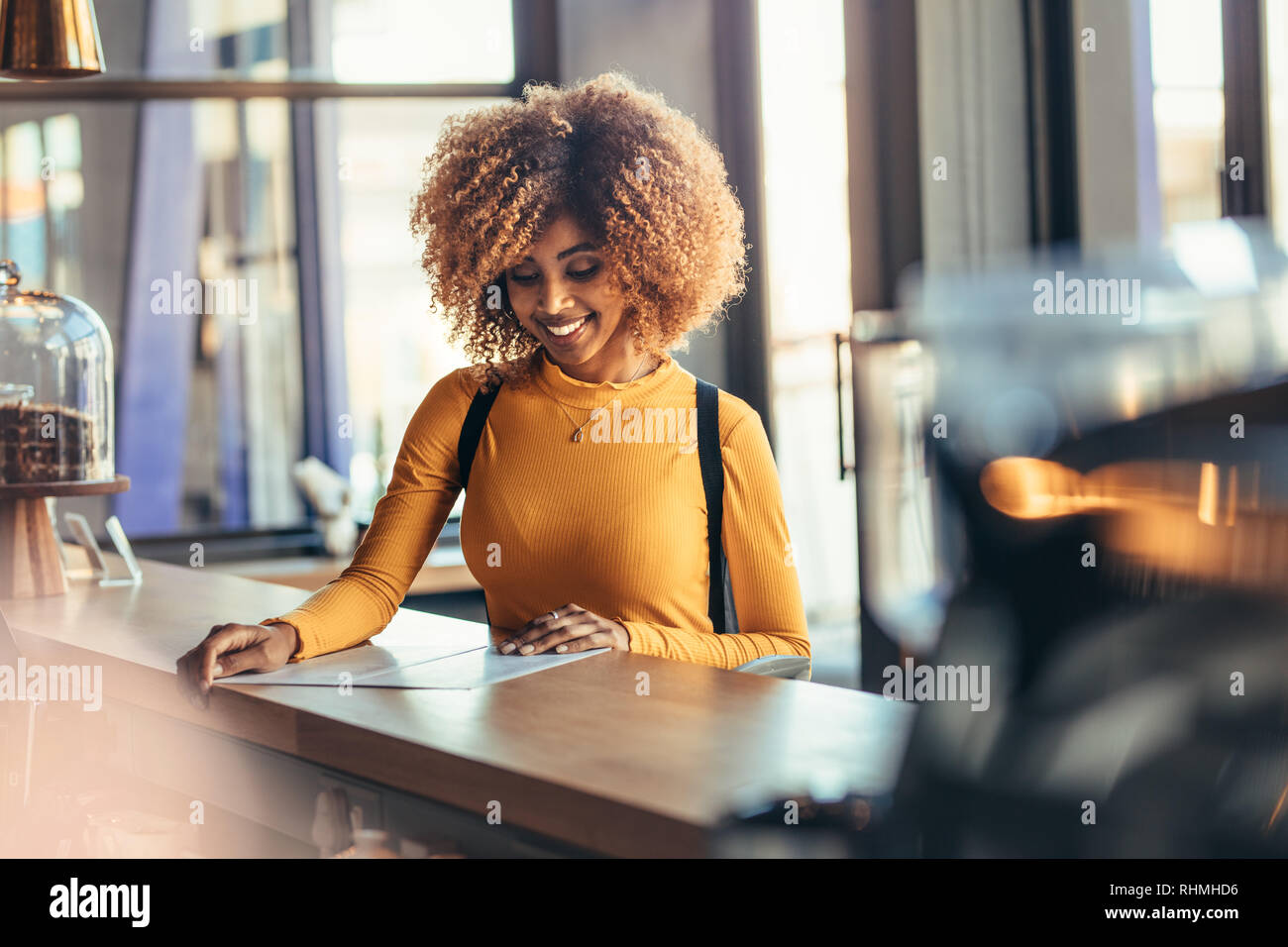 Smiling afro american woman standing at the billing counter of a coffee shop looking at the menu. Cheerful afro american traveller ordering food at a Stock Photo