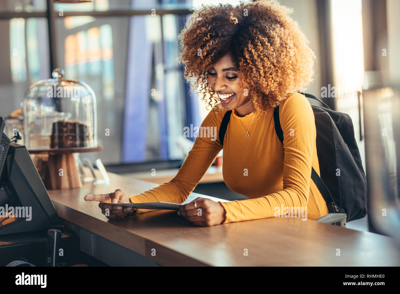 Tourist woman standing at the billing counter of a cafe holding the menu card. Smiling tourist wearing backpack talking to the cafe owner looking at t Stock Photo