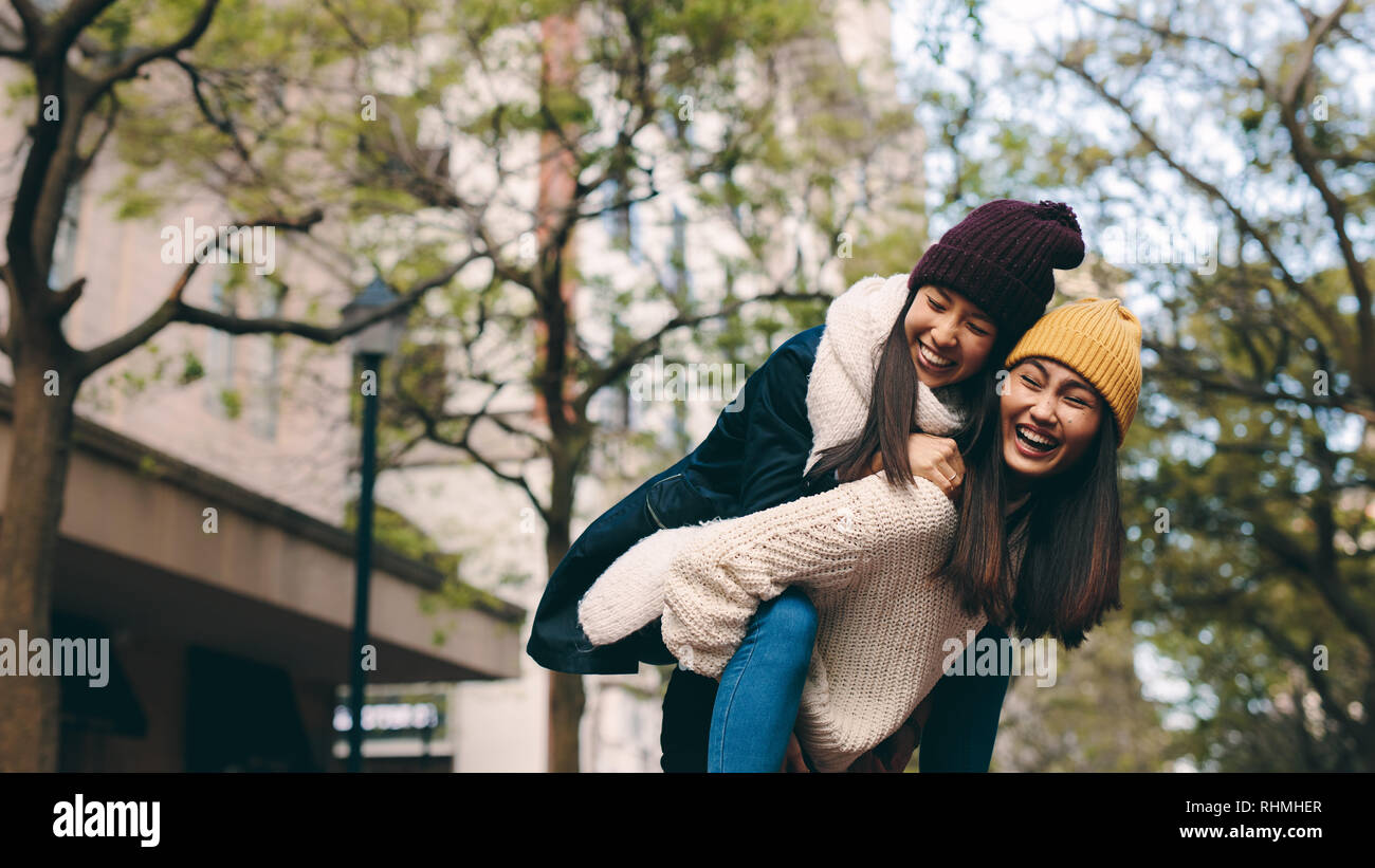 Smiling asian women in warm winter clothes playing together and having fun. Young woman piggy riding on the back of her friend on a winter morning. Stock Photo