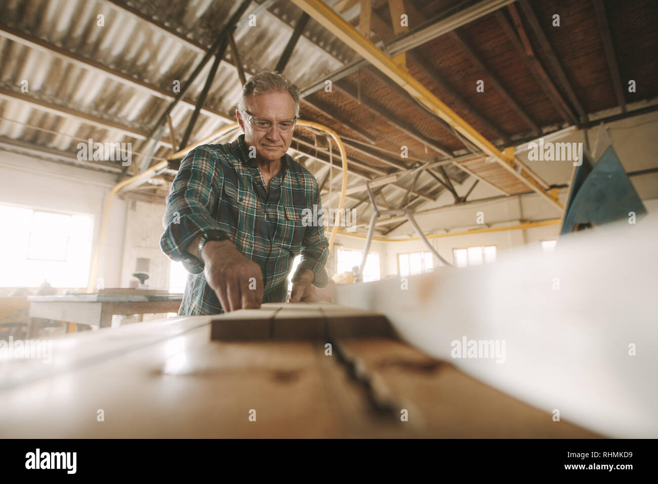 Senior male carpenter cutting wood on table saw machine. Mature caucasian man working in carpentry workshop. Stock Photo