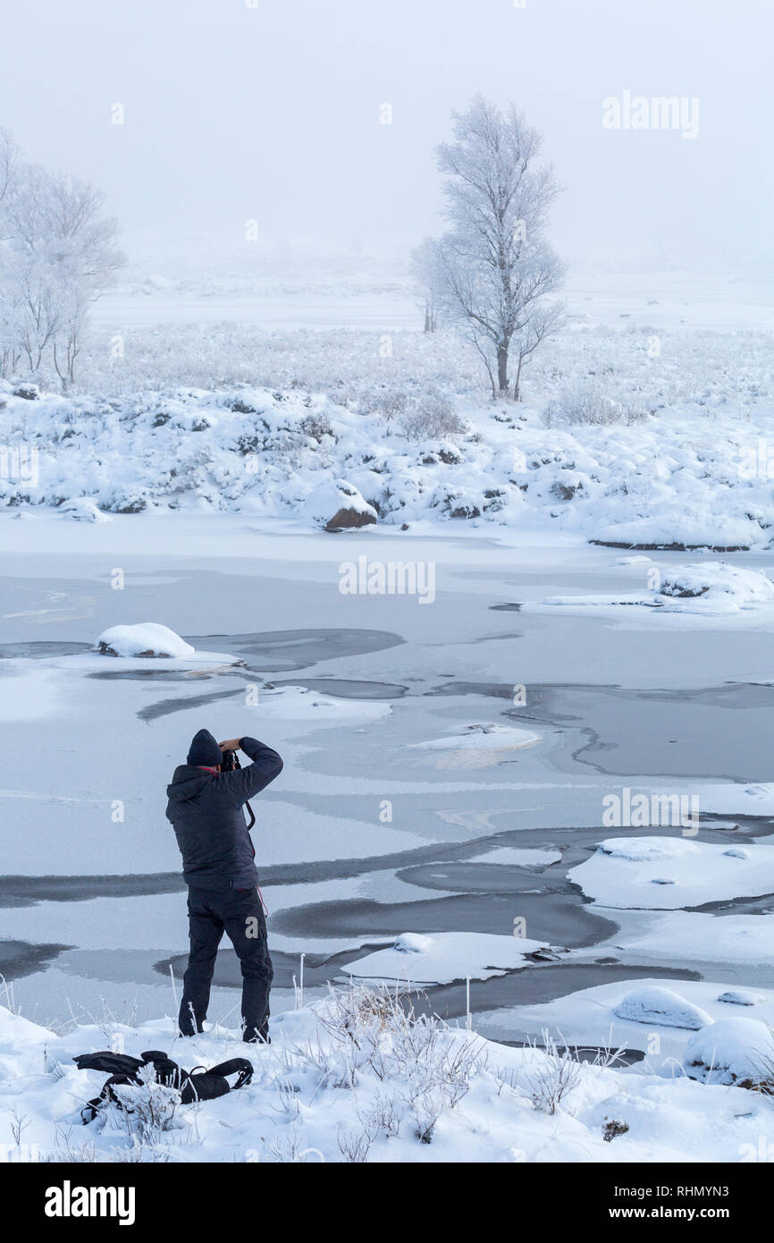 Man photographing frozen loch and tree at Loch Ba, Rannoch Moor, Scotland on a cold winter morning in January Stock Photo