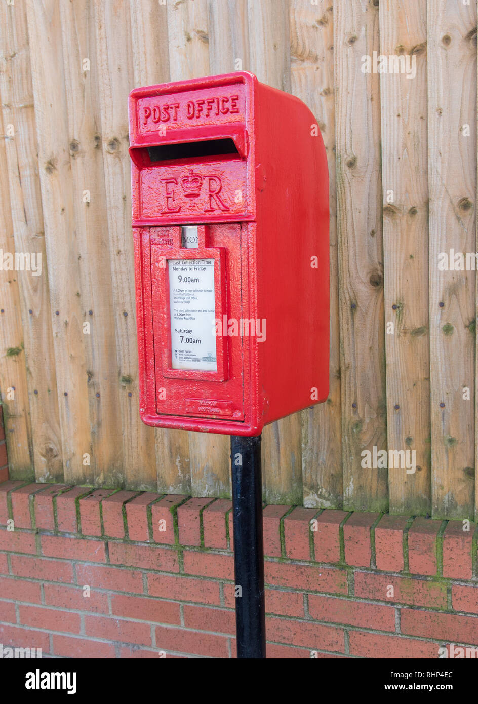 red polemounted post box showing collection times by a wooden fence Wallasey Village February 2019 Stock Photo