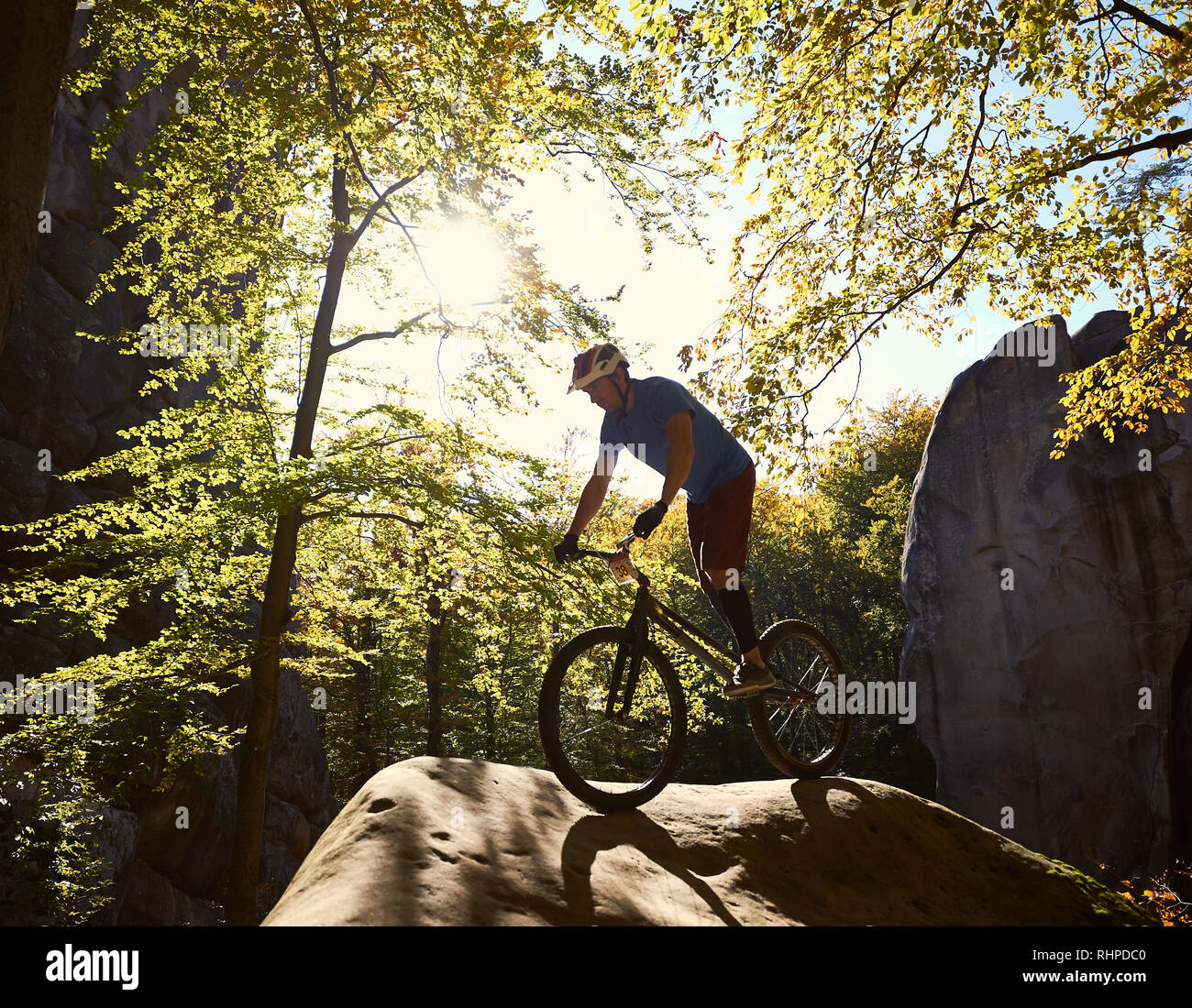 Young male cyclist balancing on trial bicycle, making acrobatic trick on big boulder in the forest on summer sunny day. Concept of extreme sport active lifestyle Stock Photo