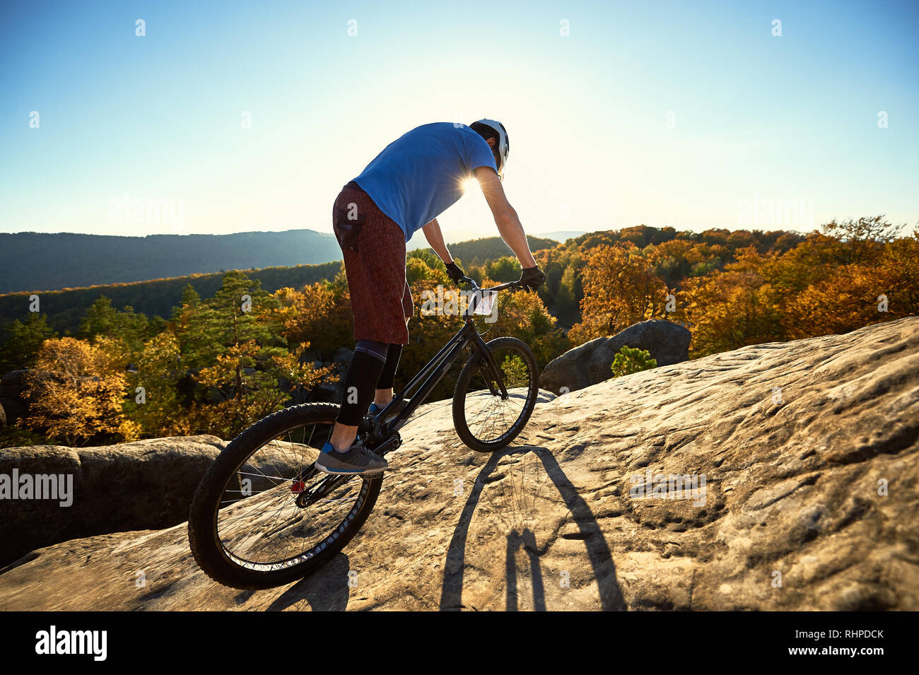Man cyclist riding on trial bike, rider making acrobatic trick on top of big boulder on summer evening, blue sky, forest and sunset on background. Concept of extreme sport active lifestyle Stock Photo