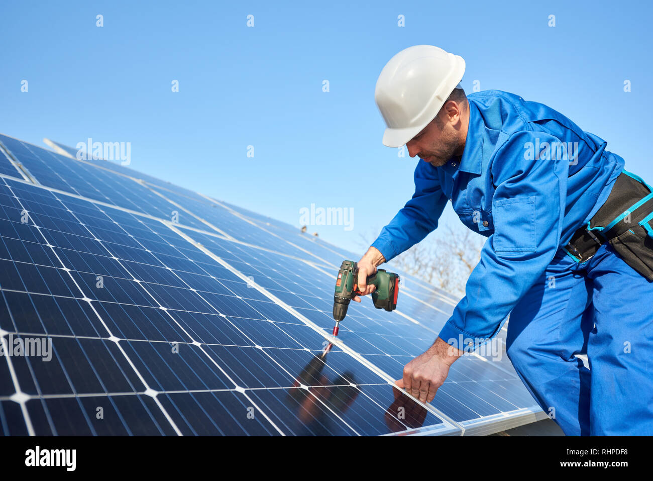 Man worker in blue suit and protective helmet installing solar photovoltaic panel system using screwdriver. Professional electrician mounting blue solar module. Alternative energy ecological concept. Stock Photo