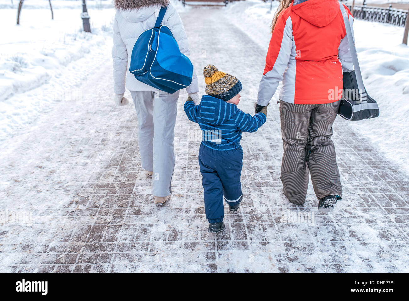 Mom and girlfriend sees the boy s son 2-6 years old by hand walking down street. Back view. Winter in city on background of snowdrifts. Returning home Stock Photo