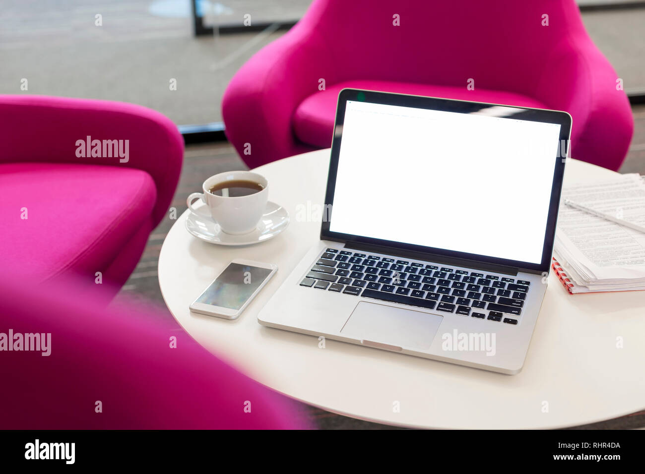 Laptop with coffee cup and smartphone on table by chairs at office Stock Photo