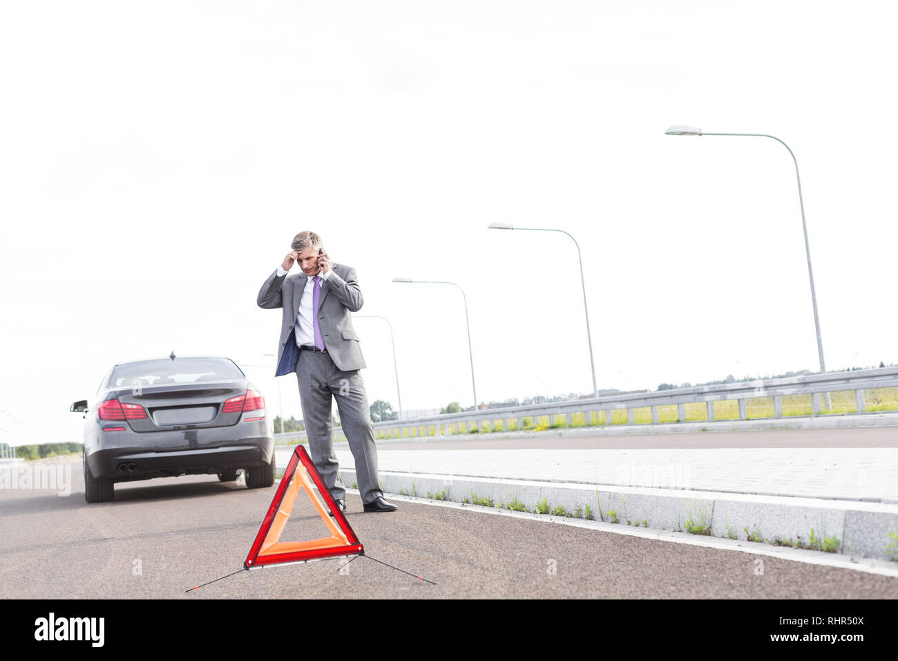 Businessman talking on phone while standing with breakdown car by warning sign on road Stock Photo