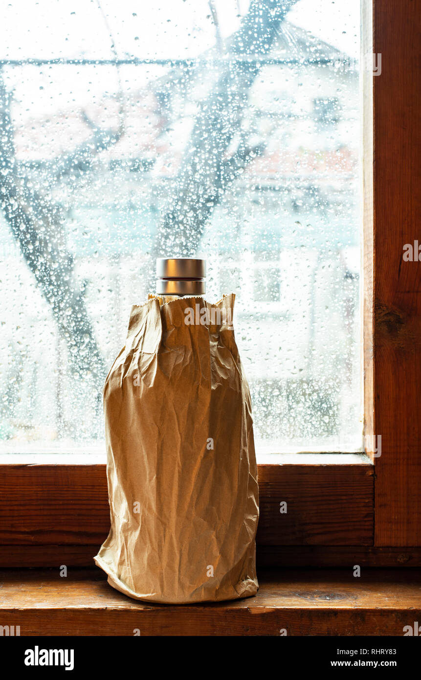 Bottle with alcohol in a brown pack paper placed to the window at rainy