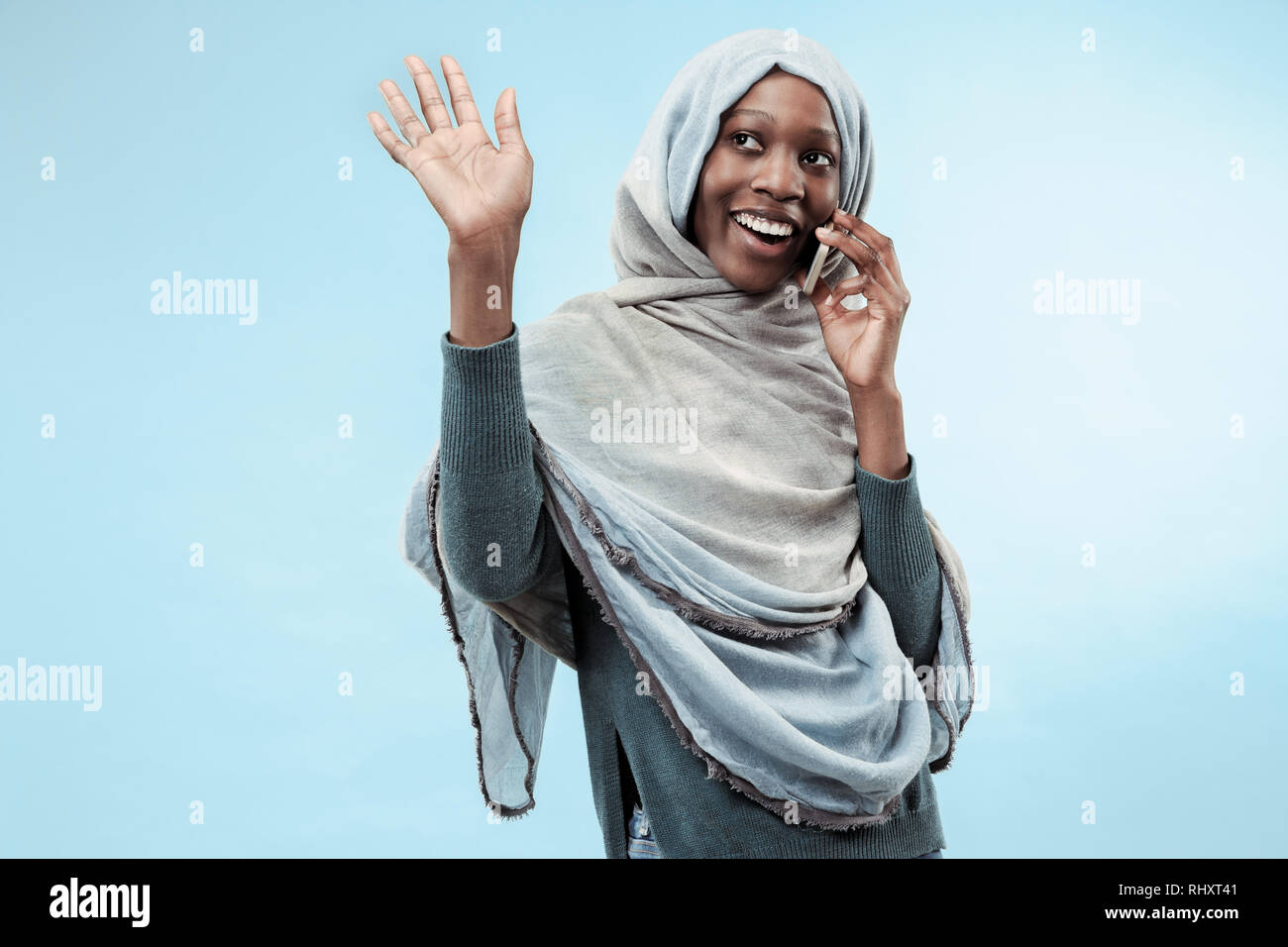 The beautiful young black african muslim girl wearing gray hijab at blue studio. She standing with a happy smile on her face. The human emotions, facial expression concept. Trendy colors Stock Photo