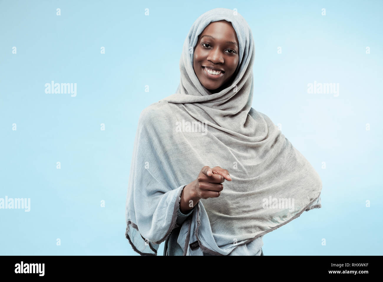 The beautiful young black african muslim girl wearing gray hijab at blue studio. She standing with a happy smile on her face. The human emotions, facial expression concept. Trendy colors Stock Photo