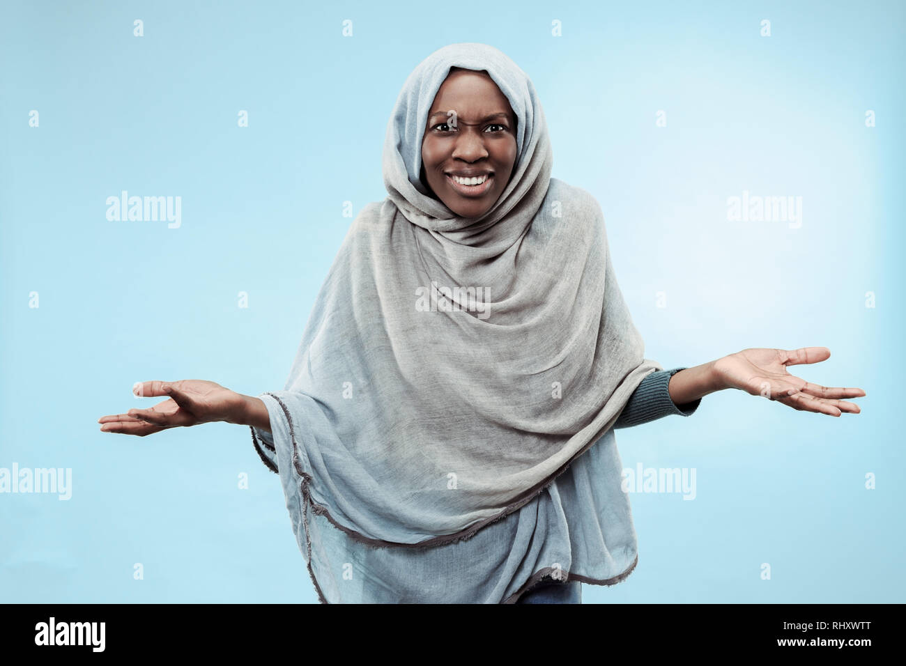 The beautiful young black african muslim girl wearing gray hijab at blue studio. She standing with a happy smile on her face. The human emotions, facial expression concept. Trendy colors Stock Photo