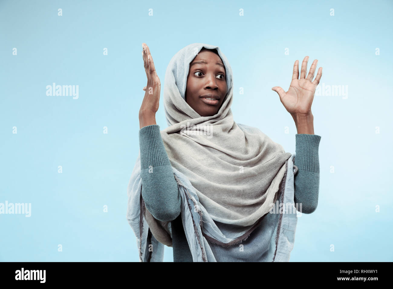 The beautiful young black african muslim girl wearing gray hijab at blue studio. She standing with a suprised emotion on her face. The human emotions, facial expression concept. Trendy colors Stock Photo