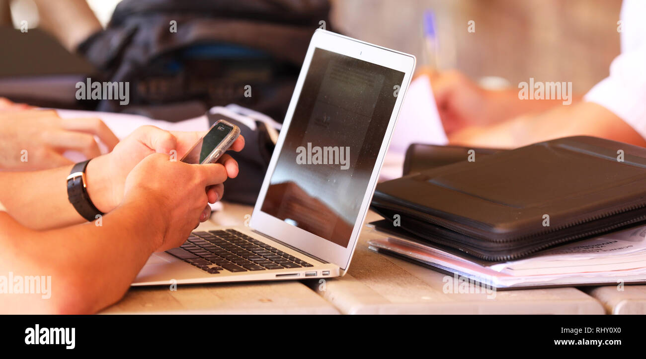 Close up of a high school student at a table or desk on a mobile cell phone texting while studying. Background of computer laptop screen and books. Stock Photo