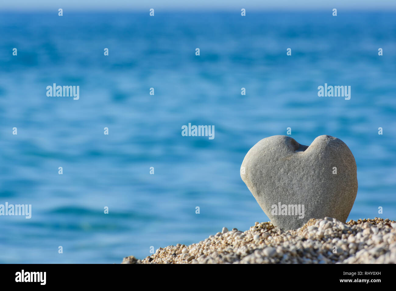 Big, grey heart shapped stone in a pebble on a beach against blurred sea and sky background, good as copy space Stock Photo