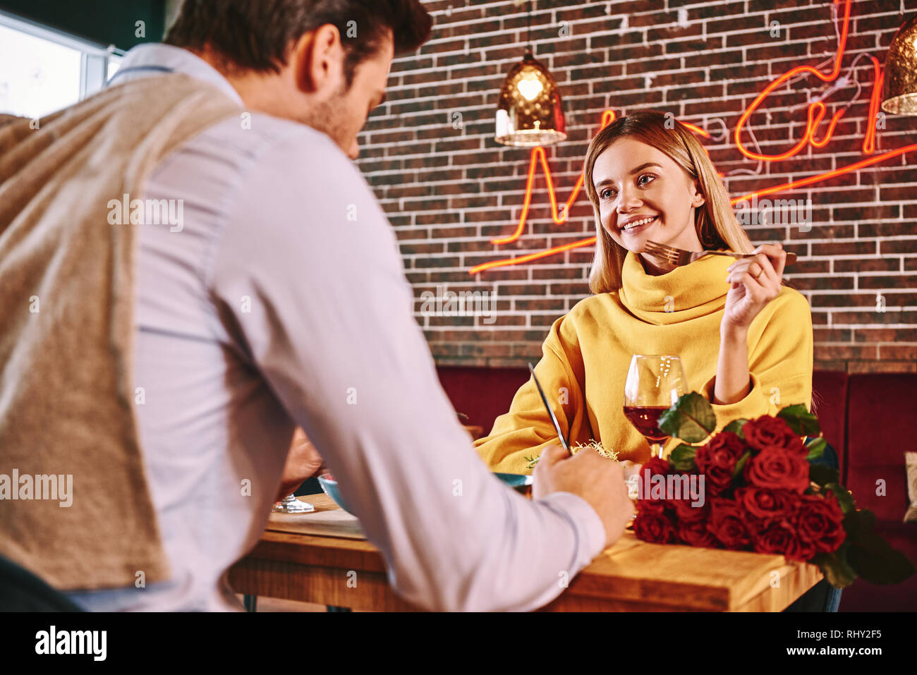 Romantic date. Blonde woman looks at her boyfriend while eating in restaurant. Red roses are lying on the table. Blonde young woman in mustard sweater Stock Photo