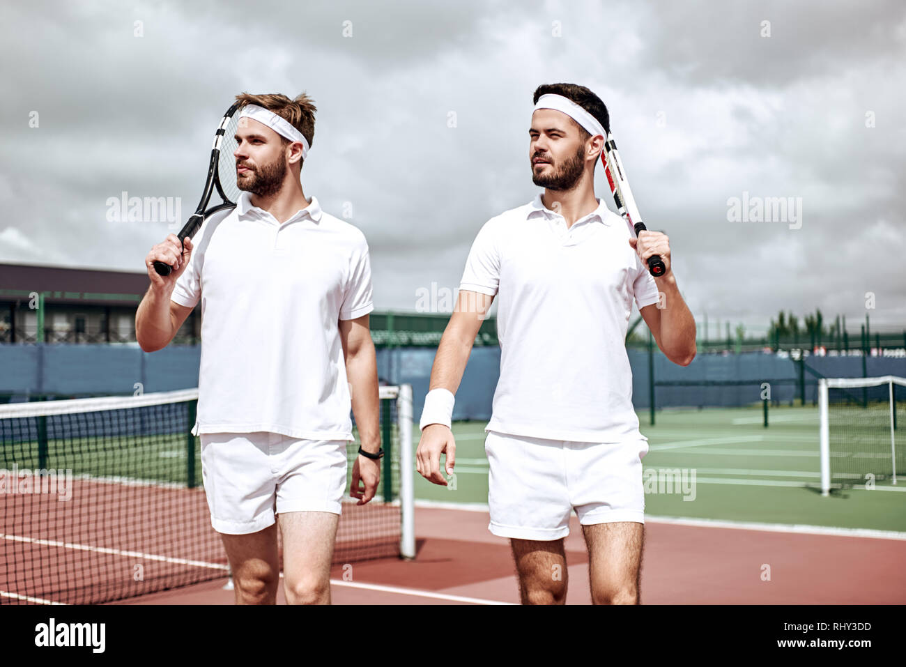 Two handsome men in white polo looking away with rackets in their hands ...