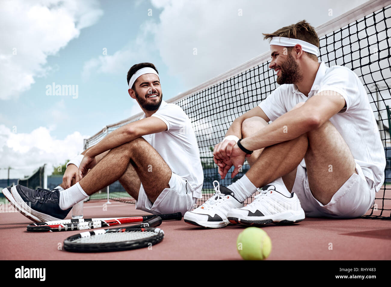 Two handsome tennis players are sitting on the court after the match, talking and smiling to each other Stock Photo